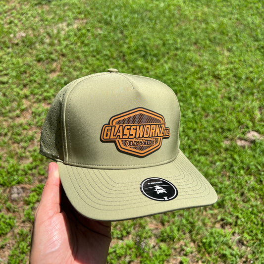 A person's hand holding an olive green trucker hat with a 'Glassworks' patch on the front and a circular black logo patch on the side, set against a background of green grass.