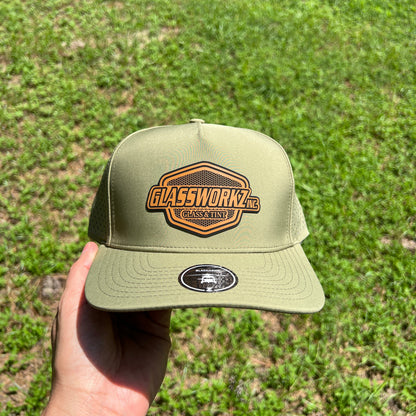 An olive green trucker hat with a brown 'Glassworkz' patch on the front, held by a hand against a background of green grass.