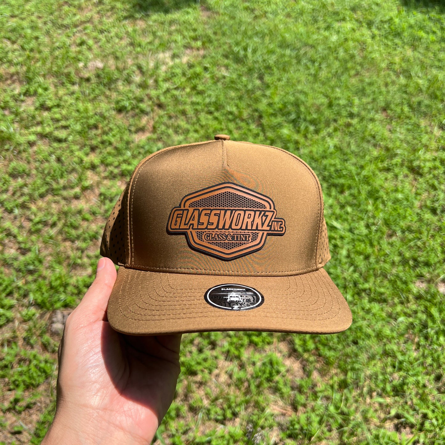 A tan trucker-style cap with a brown leather patch logo and a small oval sticker on the side, held up by a hand against a green grass background.