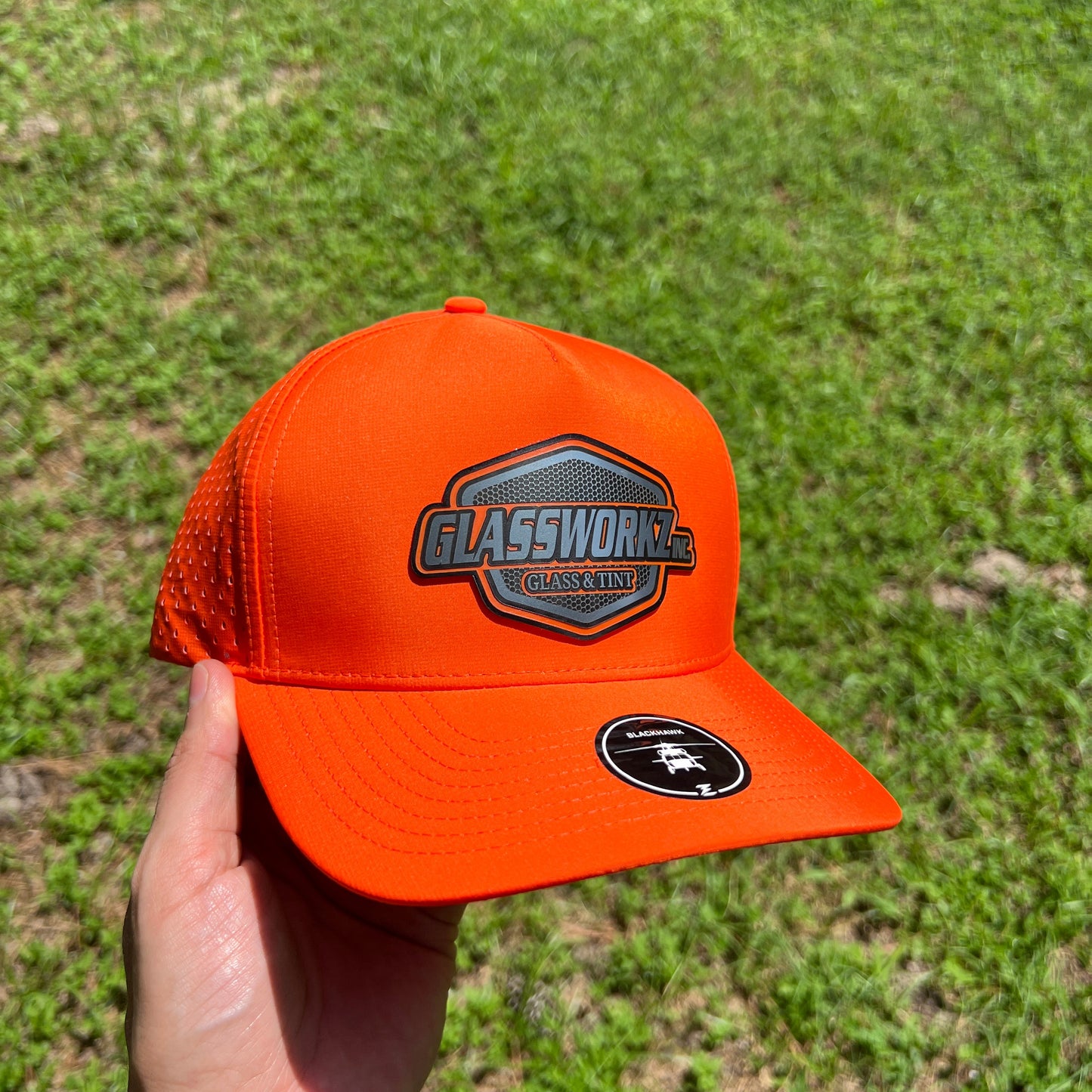An orange trucker hat with a 'GLASSWORKZ INC GLASS AND TINT' patch on the front and a circular black logo on the side, held in a hand against a background of green grass.