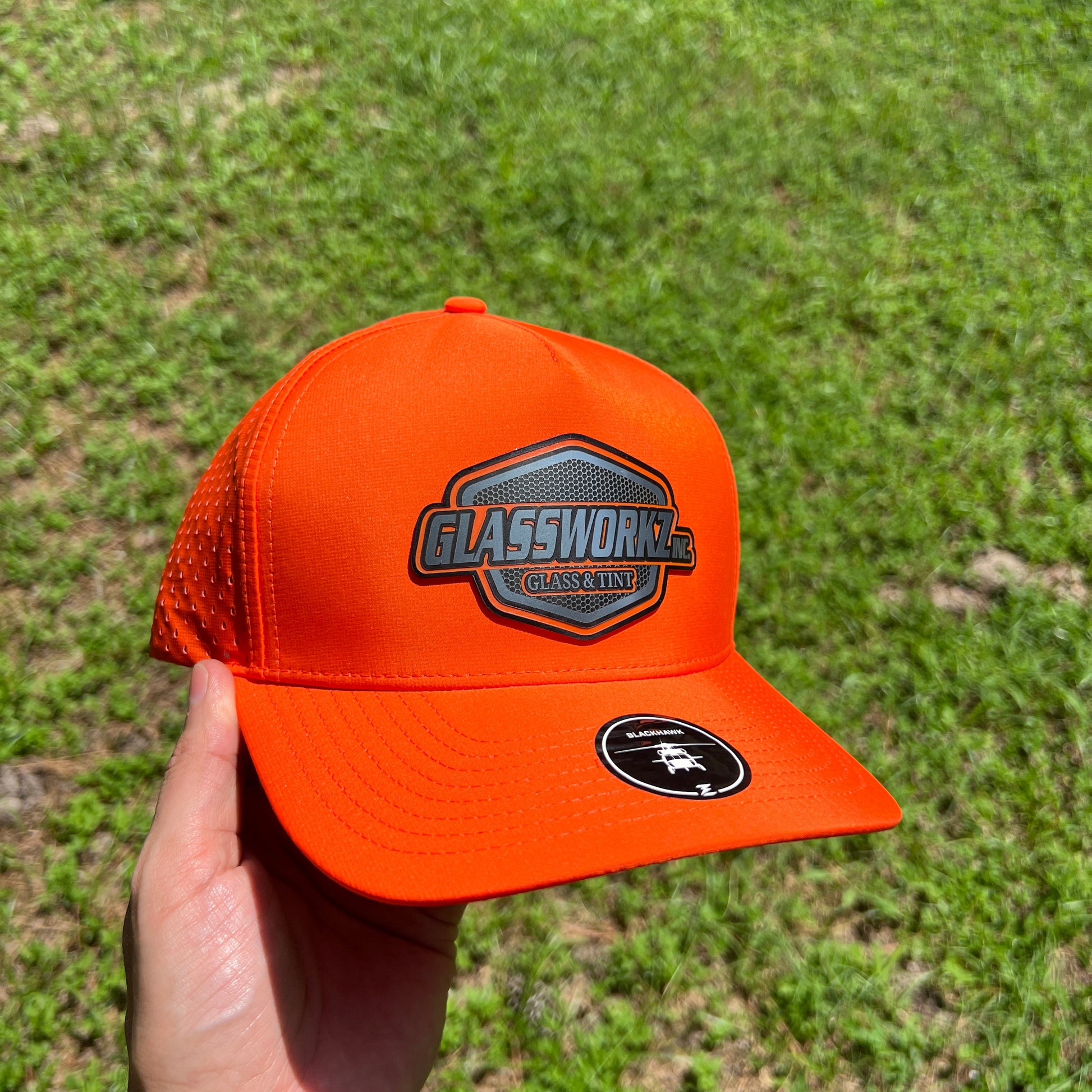An orange trucker hat with a 'GLASSWORKZ INC GLASS AND TINT' patch on the front and a circular black logo on the side, held in a hand against a background of green grass.
