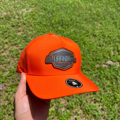 An orange trucker hat with a 'GLASSWORKZ INC GLASS AND TINT' patch on the front and a circular black logo on the side, held in a hand against a background of green grass.