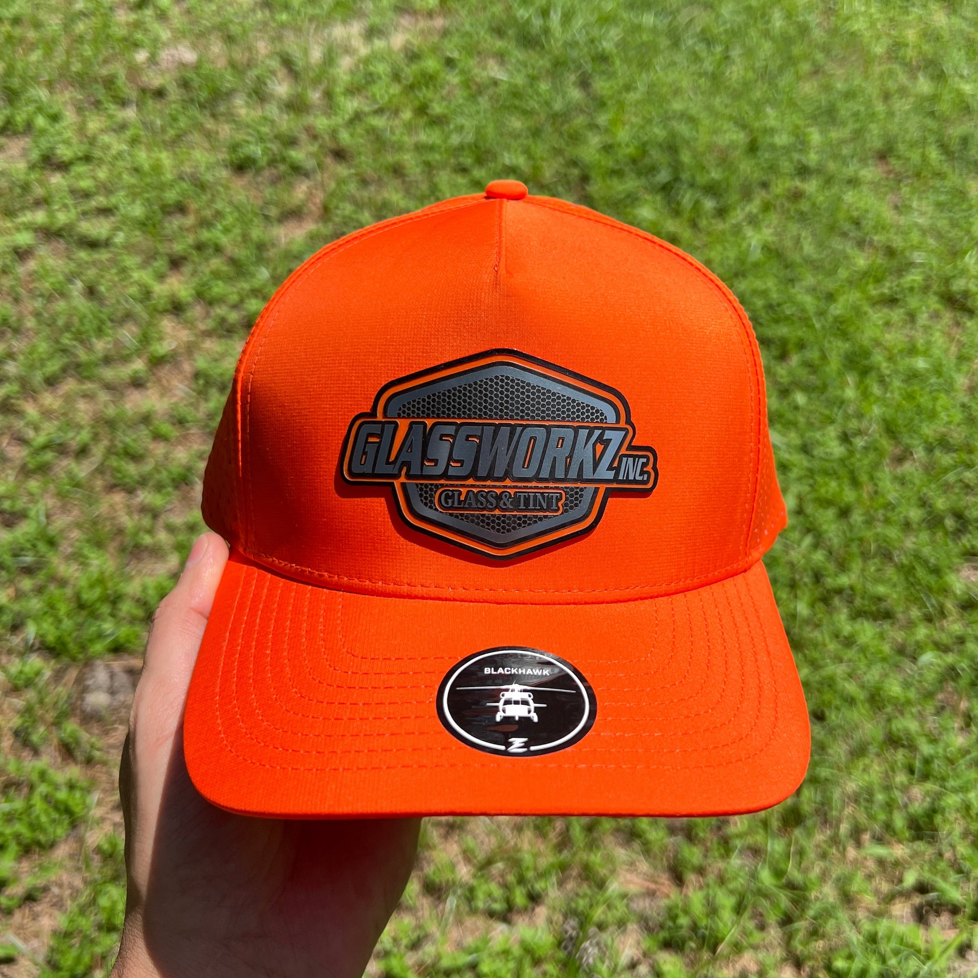 A hand holding a bright orange snapback cap outdoors with a grassy background. The cap features a black and gray hexagonal patch on the front that reads 'GLASSWORKZ INC. GLASS & TINT' and a circular black sticker on the brim.