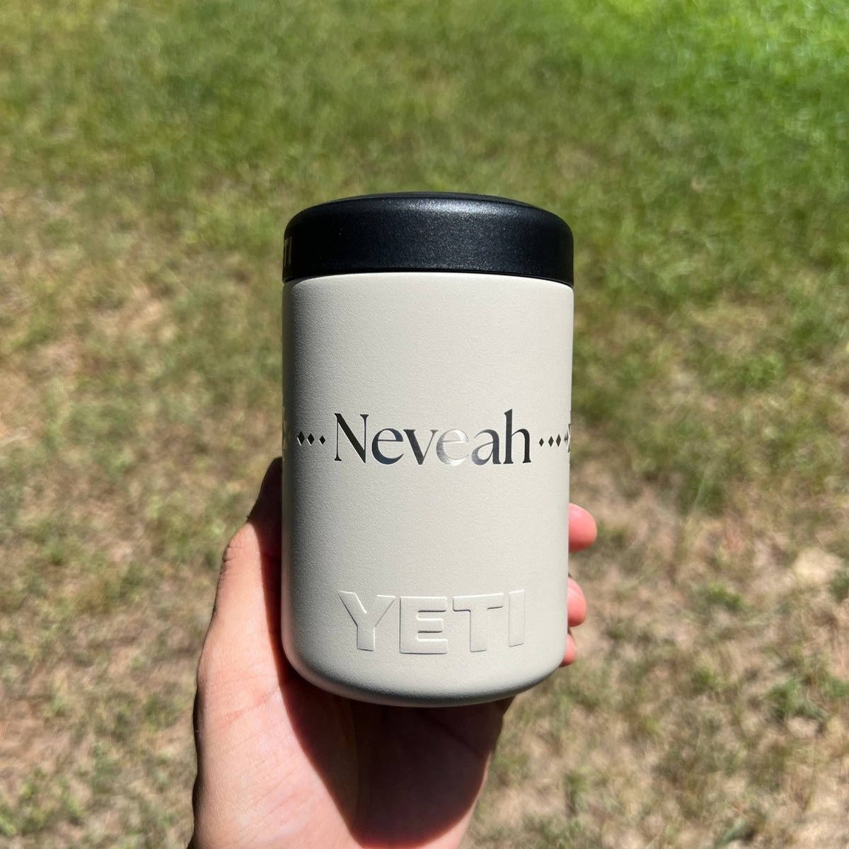 A hand holding a light gray YETI Rambler tumbler with a black lid outdoors on a grassy lawn. The tumbler features the name 'Neveah' printed in black with decorative dots on the front, and the YETI logo embossed near the bottom.