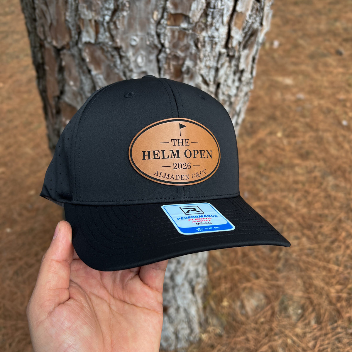 A black baseball cap held in a hand in front of a tree trunk outdoors. The cap has a tan oval patch reading “THE HELM OPEN 2026 ALMADEN GOLF” and a blue Performance Fit sticker on the brim.