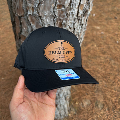 A black baseball cap held in a hand in front of a tree trunk outdoors. The cap has a tan oval patch reading “THE HELM OPEN 2026 ALMADEN GOLF” and a blue Performance Fit sticker on the brim.