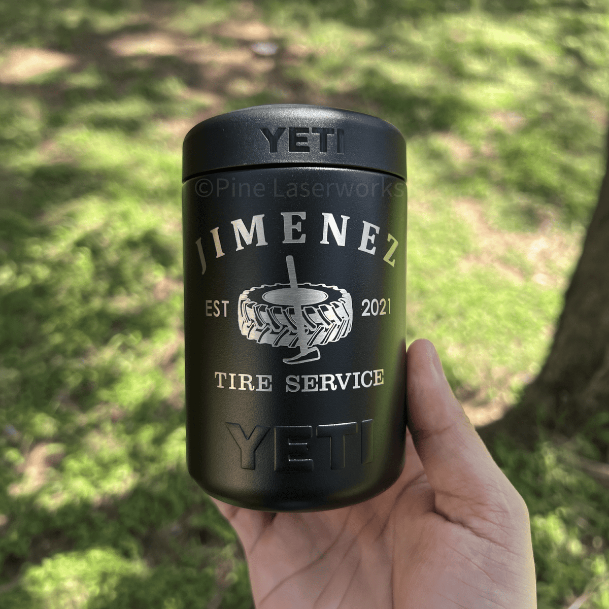 A hand holding a black YETI insulated tumbler outdoors in a grassy, sunlit area. The tumbler is engraved with 'JIMENEZ', a tire graphic, 'EST 2021', and 'TIRE SERVICE', with 'YETI' embossed at the top and bottom.