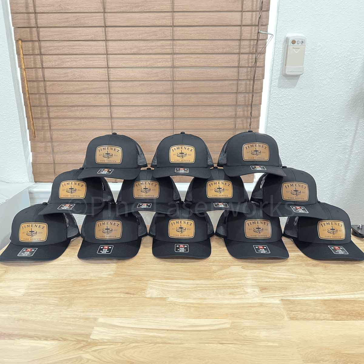 A stack of black baseball caps arranged in three rows on a light wood table, with a wooden slat door and white wall in the background. Each cap has a tan rectangular patch on the front with the word 'TIMELY' and a baseball player graphic, plus a small red and white logo on the side.