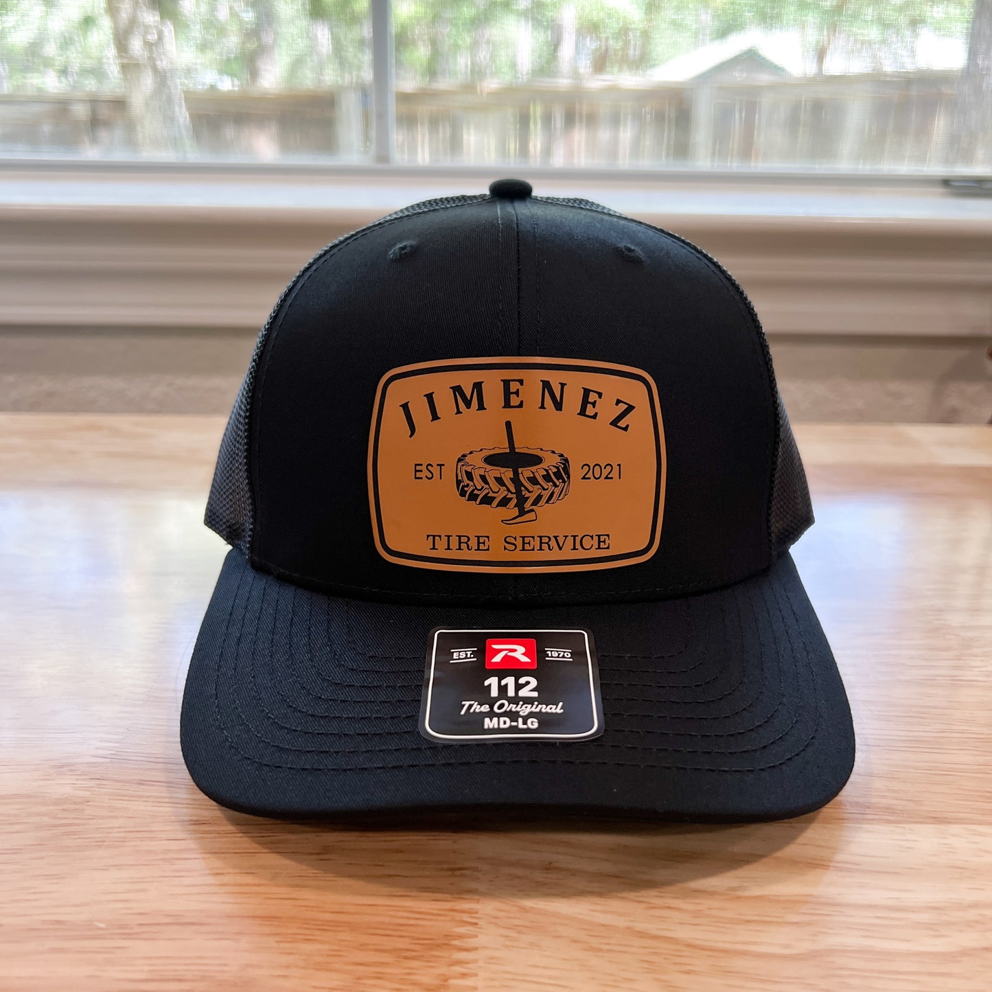 A black trucker-style cap with a tan front patch reading 'JIMENEZ EST 2021 TIRE SERVICE' and a size sticker for size 112, placed on a wooden table in front of a window.