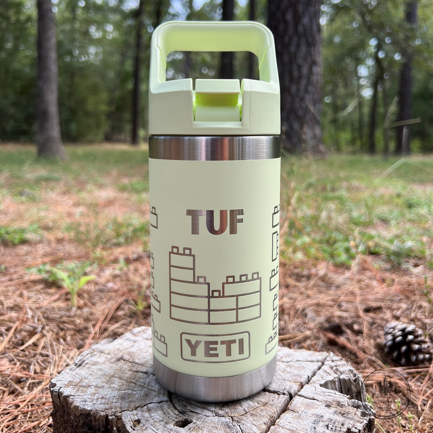 A pale yellow-green YETI TUF water bottle with a stainless steel band and a matching lid, standing on a tree stump in an outdoor forest setting.