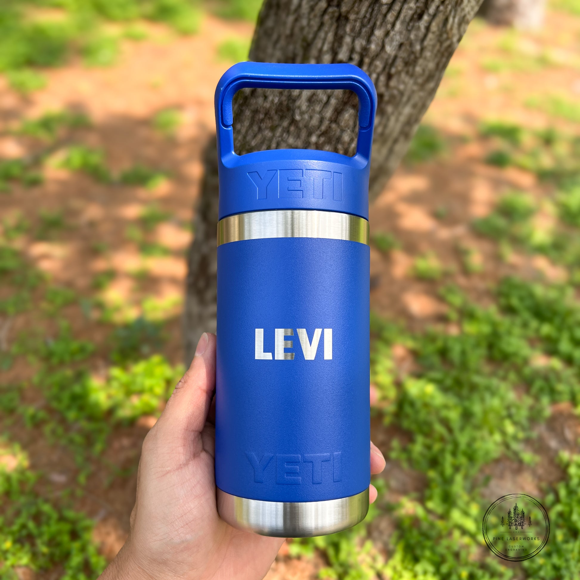 A hand holding a blue YETI insulated water bottle with a stainless steel band and the name 'LEVI' printed in white, set against a blurred outdoor background of a tree trunk and green grass.