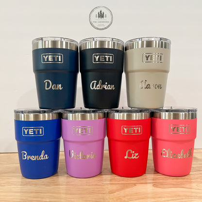 7 insulated stainless steel tumblers with clear lids are arranged on a light wood surface against a plain light wall. The tumblers come in navy, black, gray, blue, lavender, red, and pink, each with a silver YETI logo and a custom name engraved in white script.