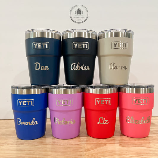 7 insulated stainless steel tumblers with clear lids are arranged on a light wood surface against a plain light wall. The tumblers come in navy, black, gray, blue, lavender, red, and pink, each with a silver YETI logo and a custom name engraved in white script.