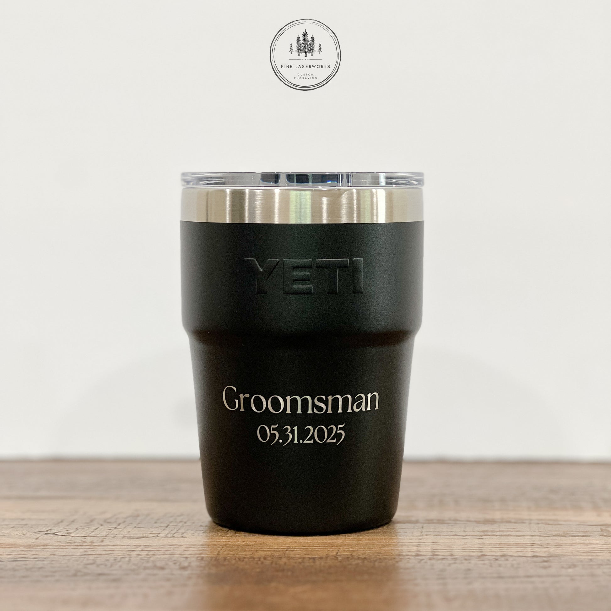 A black YETI insulated tumbler with a clear lid sits on a wooden surface against a light background. The tumbler is engraved with the word 'Groomsman' and the date '05.