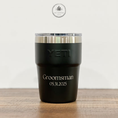 A black YETI insulated tumbler with a clear lid sits on a wooden surface against a light background. The tumbler is engraved with the word 'Groomsman' and the date '05.