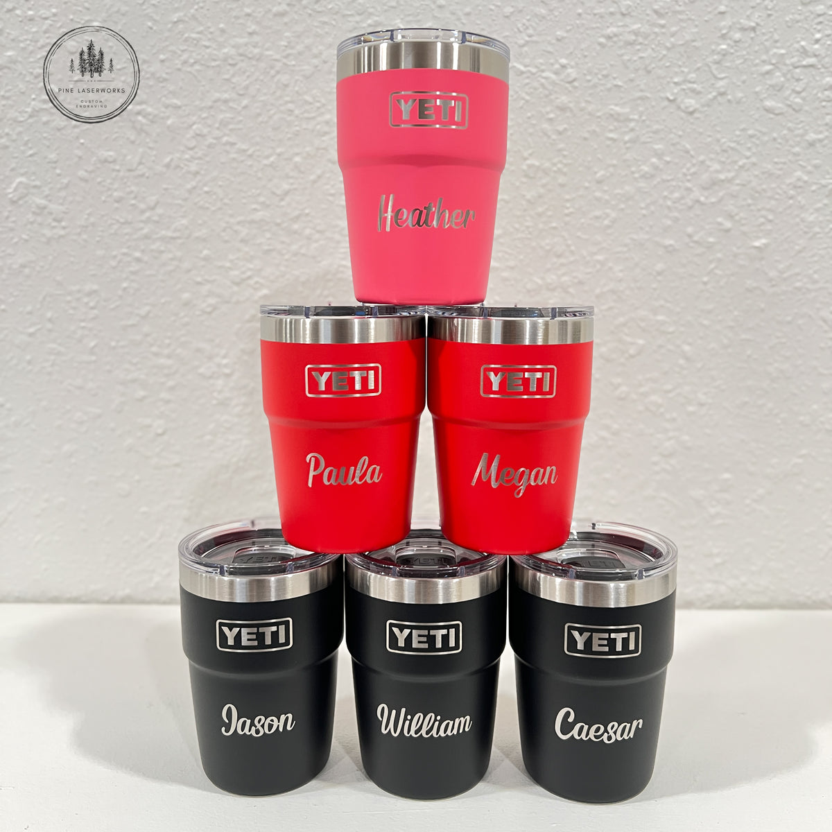 A pyramid arrangement of seven customized YETI travel tumblers on a white surface against a textured white wall. The top of the pyramid features a single pink tumbler, followed by two red tumblers, then four black tumblers at the base.