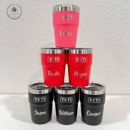 A pyramid arrangement of seven customized YETI travel tumblers on a white surface against a textured white wall. The top of the pyramid features a single pink tumbler, followed by two red tumblers, then four black tumblers at the base.