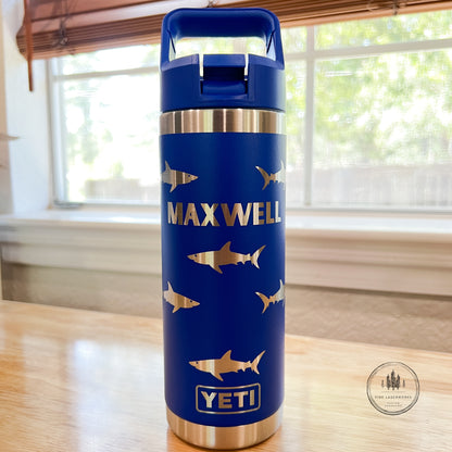 A blue YETI insulated water bottle with gold shark silhouettes and the name 'MAXWELL' printed on the front, sitting on a wooden table in front of a window.