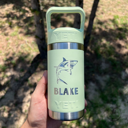 A hand holding a grasshopper YETI insulated water bottle outdoors. The bottle features a shark graphic and the name 'BLAKE' printed on the front, with stainless steel accents at the top and bottom.