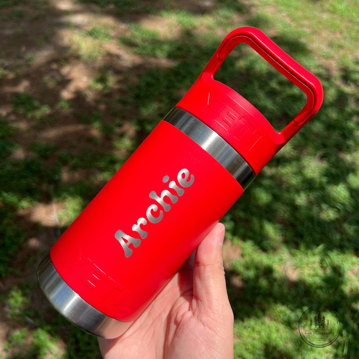 A hand holding a bright red insulated stainless steel tumbler with a matching red carrying loop lid. The word 'Archie' is printed in silver on the side, and the background is a blurred outdoor grassy area.