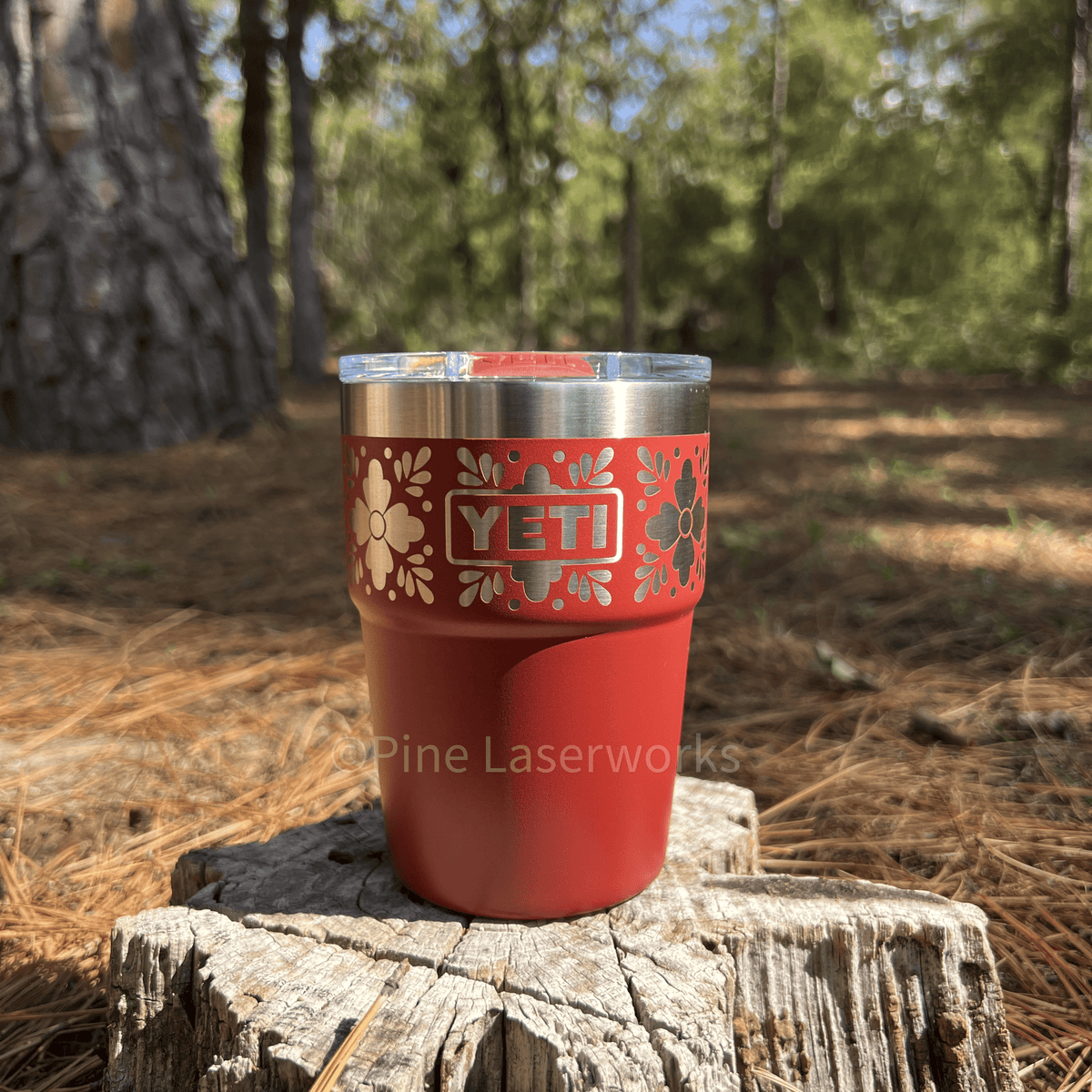 A red YETI insulated tumbler with a clear lid and silver rim sits on a tree stump in an outdoor forest setting with pine needles and trees in the background.