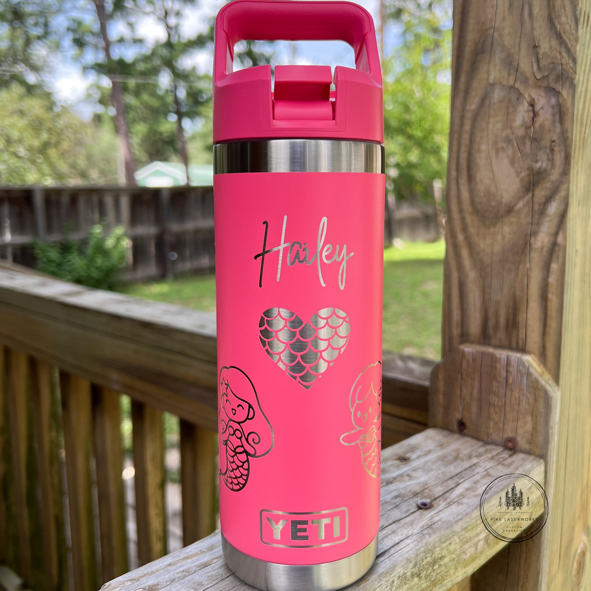 A pink YETI water bottle with a matching pink lid, personalized with the name 'Harley' and mermaid-themed graphics, sitting on a wooden deck railing with a blurred outdoor yard in the background.