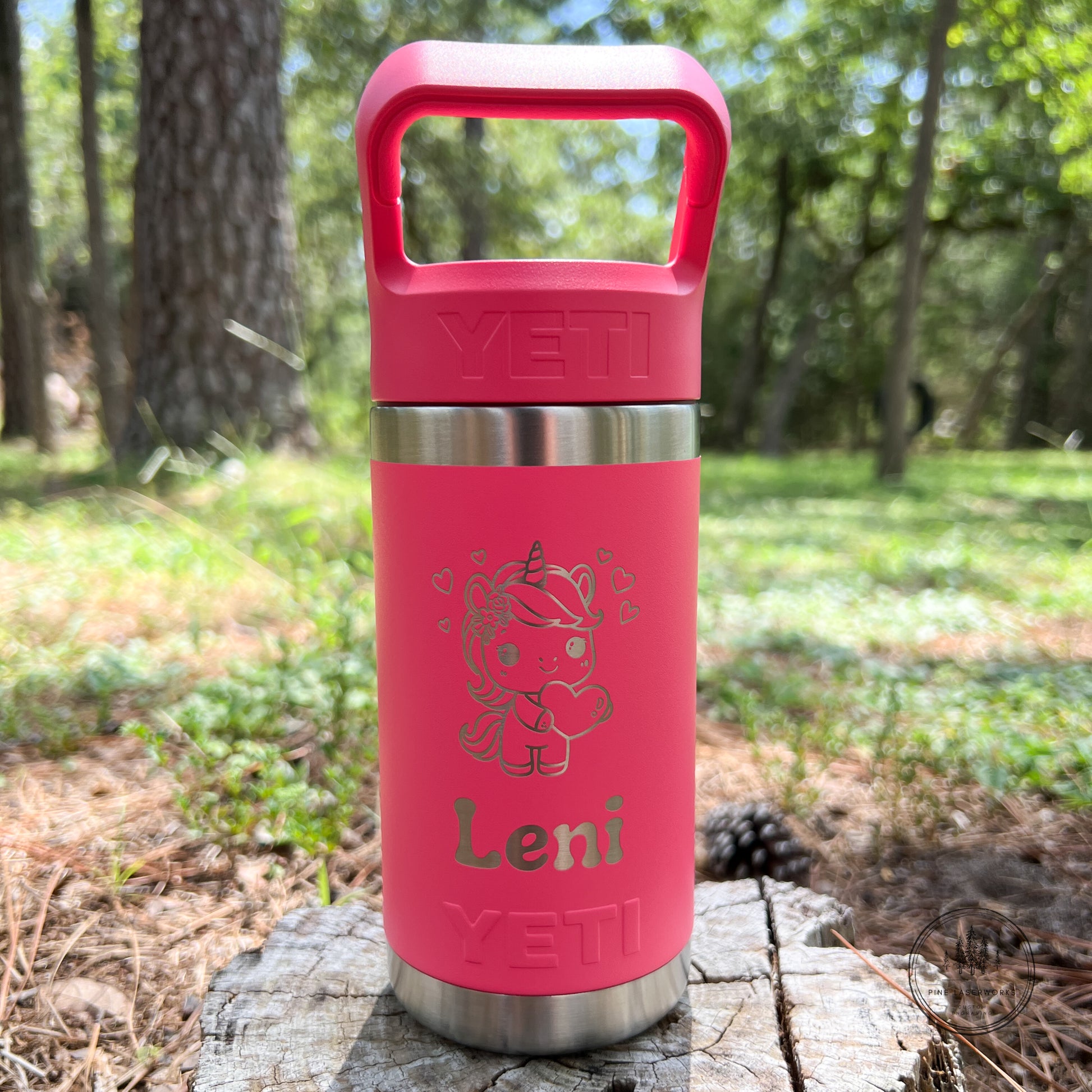 A pink YETI insulated tumbler with a matching pink lid and handle, standing on a tree stump in a forest setting. The tumbler features a silver stainless steel band, a custom cartoon character graphic, and the name 'Leni' printed in silver.