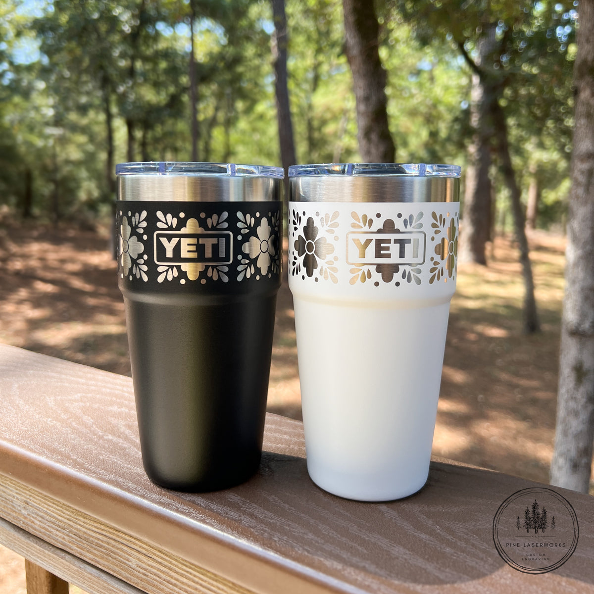 Two YETI insulated tumblers with floral laser-cut bands and clear lids, shown side by side on a wooden railing outdoors with trees in the background. One tumbler is matte black and the other is matte white.
